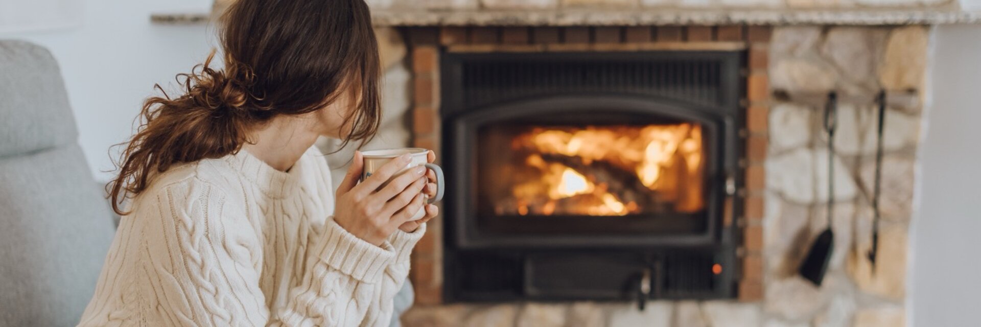 Homeowner warming up with a mug beside a wood-burning insert in a stone fireplace