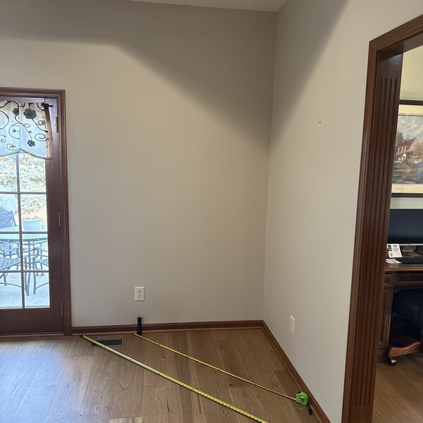 Before—bare two-story great-room wall with tape measure on the floor mid-consultation